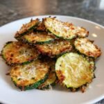A pile of crispy, golden-brown Garlic Parmesan Baked Zucchini Chips topped with shredded Parmesan cheese on a white plate.
