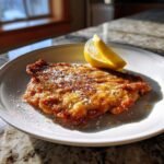 A golden brown, pan-fried Chicken Paillard dusted with powdered sugar, served with lemon wedges on a white plate.