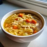 A close-up of a white bowl filled with steaming Chicken Noodle Soup, featuring shredded chicken, egg noodles, and bright carrots.