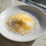 A small pile of white sugar mixed with bright yellow lemon zest to make Lemon Sugar on a white saucer.