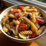 A close-up view of a vibrant bowl of Italian Pasta Salad featuring rotini pasta, cherry tomatoes, cucumbers, red onion, and olives.