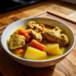 A close-up of a white bowl filled with Jamaican Curry Chicken, featuring chunks of yellow-sauced chicken, potatoes, and bright orange carrots.