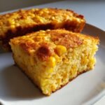 Close-up of a square slice of moist Cornbread Casserole studded with whole corn kernels on a white plate.