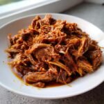 A close-up of tender, shredded Braised Pork Shoulder coated in rich, dark sauce on a white plate.