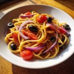 Close-up of a bowl of colorful Spaghetti Salad featuring cherry tomatoes, black olives, and bright red onion slices.