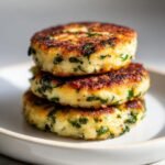 A stack of three golden-brown Spinach Garlic Potato Patties, flecked with bright green herbs, served on a white plate.