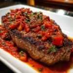 A thick, seared steak topped generously with chunky tomato sauce and fresh herbs, served on a white plate, showcasing Steak Pizzaiola.