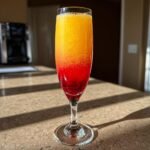 A vibrant, layered Sunrise Mimosa cocktail showing red, orange, and yellow gradients in a champagne flute glass.