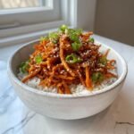 A bowl of white rice topped with shredded, saucy sweet potato rice teriyaki and garnished with sesame seeds and green onions.