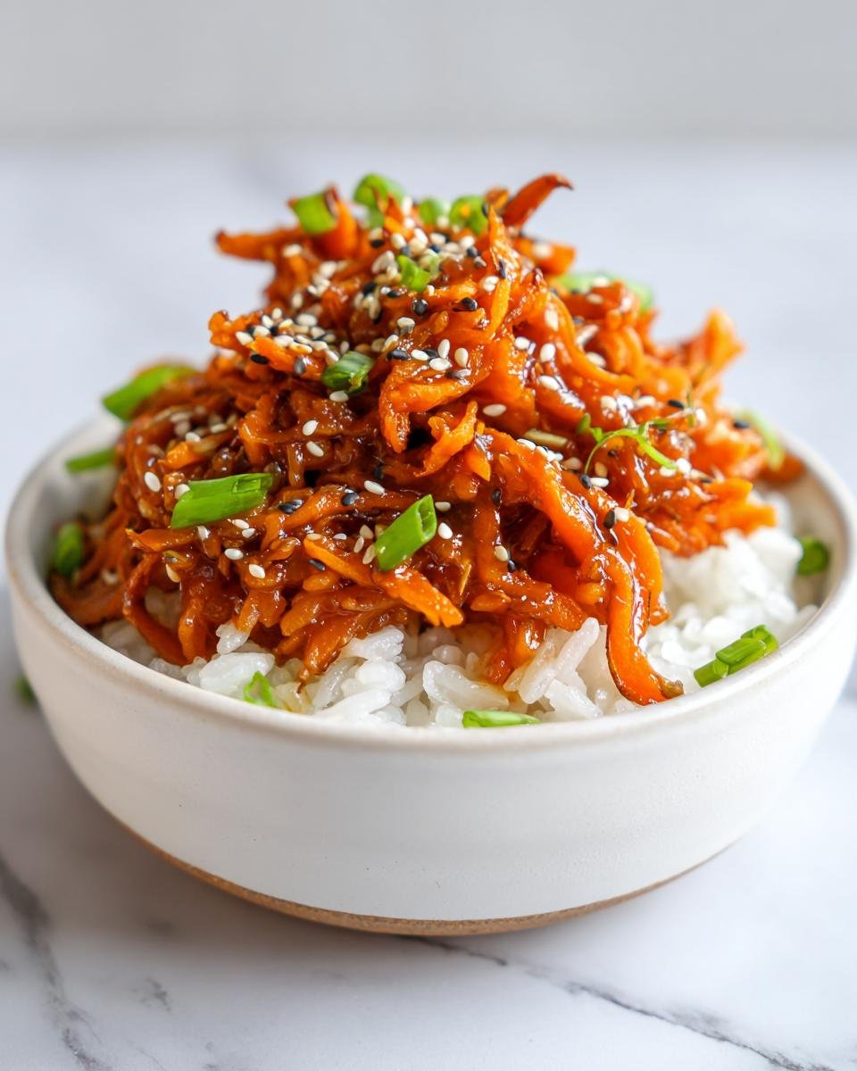 Close-up of shredded sweet potato in teriyaki sauce served over white rice, topped with sesame seeds and green onions in a white bowl.