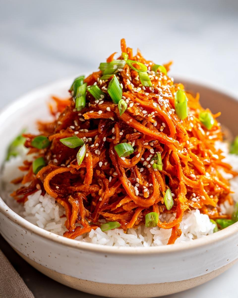 Close-up of shredded sweet potato coated in teriyaki sauce served over white rice, topped with sesame seeds and green onions.