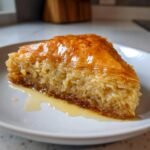 A single, glistening slice of Coconut Baklava resting on a white plate with syrup pooled underneath.