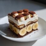 A layered slice of Banana Icebox Cake featuring cream, ladyfingers, and fresh banana slices, dusted with cocoa powder.