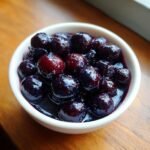 A close-up view of glossy, dark purple blueberries swimming in syrup, served as Blueberry Compote in a small white bowl.