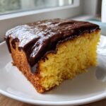 A close-up of a moist slice of yellow Boston Cream Pie Poke Cake topped with rich, dripping chocolate ganache.