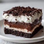 Close-up of a layered slice of Chocolate Poke Cake featuring dark cake, chocolate pudding, and whipped cream topped with crumbs.