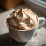 A white mug overflowing with perfectly piped Coffee Whipped Cream, catching the sunlight.