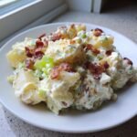 A close-up of creamy Bacon Potato Salad served on a white plate, topped with crispy bacon bits and celery.