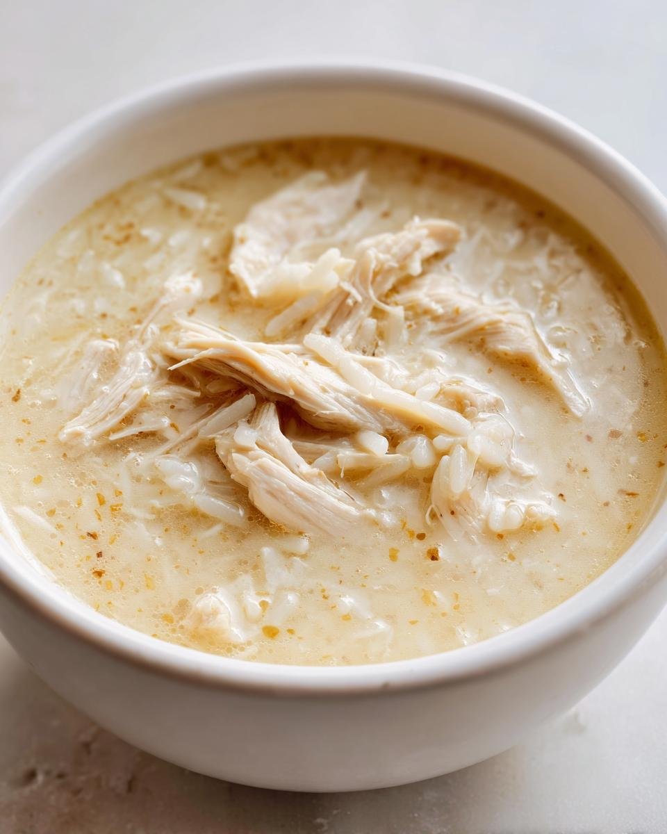 Close-up of a white bowl filled with creamy Greek Lemon Chicken Soup, featuring shredded chicken and rice.