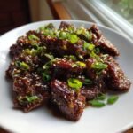 Close-up of glossy, saucy Beef Teriyaki pieces topped with green onions and sesame seeds on a white plate.