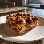 A single serving of rich, gooey Seven Layer Bars featuring oats, chocolate chips, and caramel topping, sitting on a white plate.