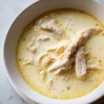 A close-up overhead view of a white bowl filled with creamy, yellow Greek Lemon Chicken Soup topped with shredded chicken.
