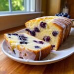 A slice of moist Lemon Blueberry Pound Cake drizzled with white lemon glaze, next to the loaf.
