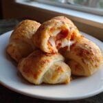A stack of golden-brown Pepperoni Pizza Crescent Rolls on a white plate, one is broken open showing melted cheese and pepperoni.