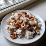A white plate piled high with Christmas Reindeer Crack snack mix, coated in powdered sugar, featuring cereal, pretzels, and pecans.