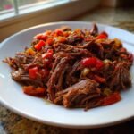 A close-up of tender, shredded Mississippi Pot Roast mixed with chunks of red and green peppers on a white plate.
