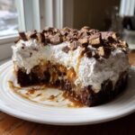 A slice removed from a rich Snickers Poke Cake showing chocolate cake, caramel filling, whipped topping, and chopped Snickers.