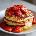 A close-up of a stack of golden brown Strawberry Pancakes topped with sliced fresh strawberries and drizzled with syrup.