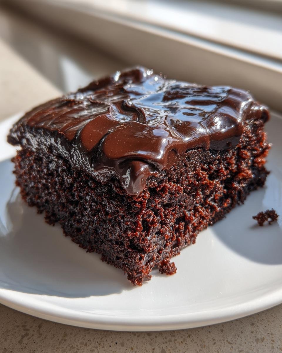 A close-up of a moist slice of Texas Sheet Cake topped with thick, glossy chocolate frosting, sitting on a white plate.