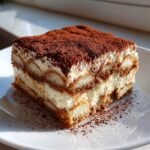 A perfect slice of Tiramisu, showcasing layers of cream and coffee-soaked ladyfingers, dusted heavily with cocoa powder, a classic Italian dessert.