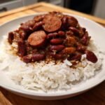 A close-up view of a bowl of white rice topped generously with savory Red Beans And Rice and sliced sausage.