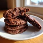 A stack of four rich, dark chocolate Fudge Rounds cookies on a white plate, one broken open to show the gooey center.