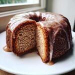 A close-up of an Apple Cider Donut Bundt Cake cut in half, showing the moist interior and dripping cinnamon glaze.