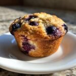 A close-up of a perfectly baked Banana Blueberry Muffins with visible blueberries on a white plate.