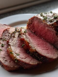 Slices of medium-rare Beef Tenderloin Roast, seasoned and garnished with fresh thyme, resting in its juices.