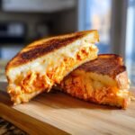 A Buffalo Chicken Grilled Cheese Sandwich cut diagonally, showing melted cheese and shredded orange buffalo chicken filling.