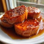 Four thick slices of glossy, glazed Candied Sweet Potatoes sitting in a rich brown syrup on a white plate.
