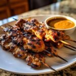 Close-up of grilled Chicken Satay With Peanut Sauce skewers on a white plate next to a small bowl of sauce.