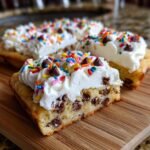 Close-up of a slice of chocolate chip Dessert Pizza topped with white frosting, sprinkles, and mini chocolate chips.