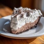 A decadent slice of Chocolate Cream Pie featuring a dark crust, chocolate filling, and tall layer of whipped cream topped with shavings.