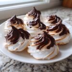 A close-up of several perfectly piped Chocolate Tipped Meringues with glossy chocolate swirls on a white plate.