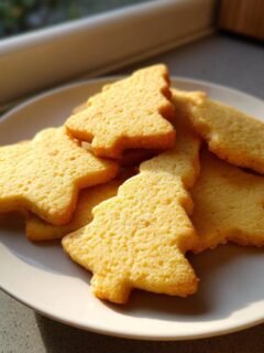 A pile of golden, star and tree-shaped Christmas Butter Cookies resting on a white plate near a window.