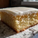A close-up, sunlit shot of a thick, square slice of Frosted Sugar Cookie Bars showing the soft yellow cake base and thick white frosting.
