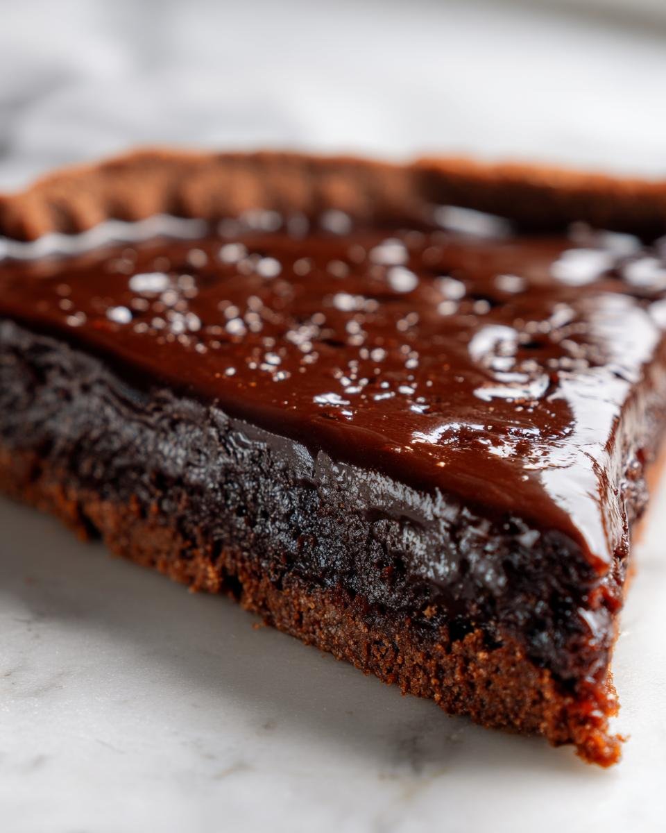 A close-up, macro shot of a rich, dark slice of Fudge Dessert Pizza showing the dense base and a shiny chocolate glaze topped with sea salt flakes.