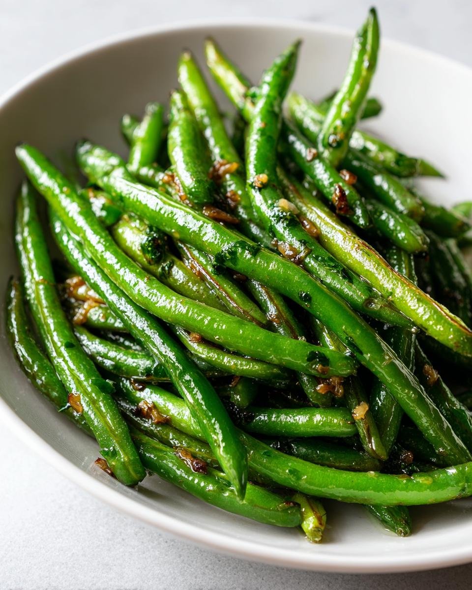 A close-up shot of vibrant, glossy Garlic Butter Green Beans tossed with browned garlic pieces in a white bowl.