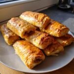 A stack of golden-brown, flaky Christmas Sausage Rolls piled high on a light grey plate.